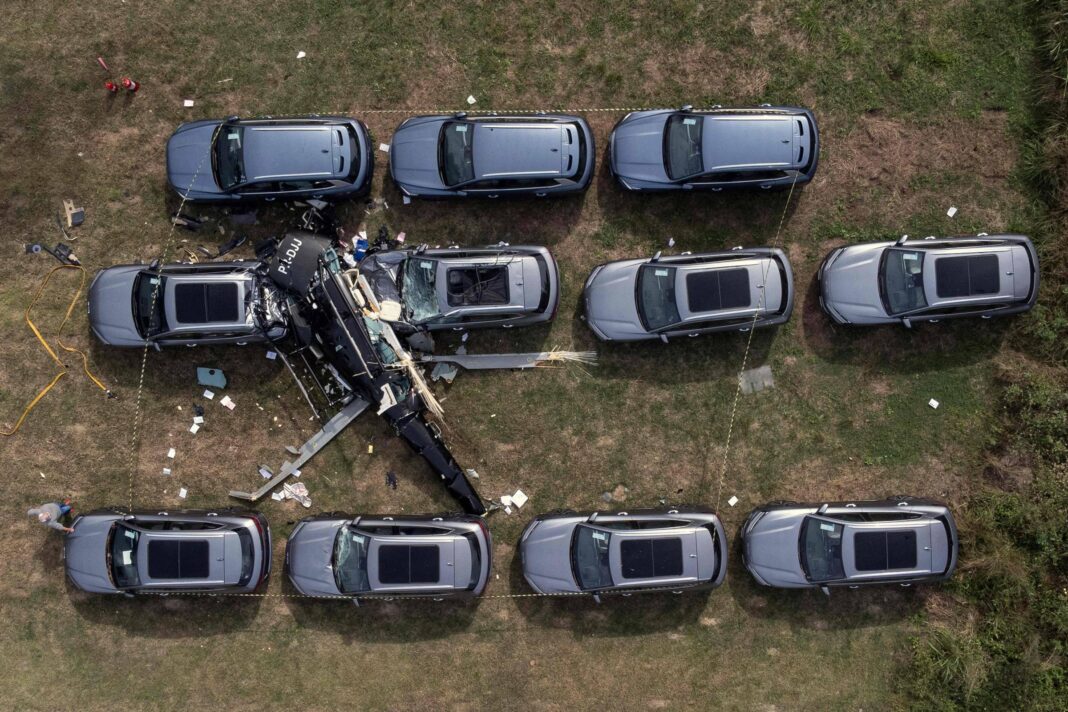 Helicóptero preto caiu entre 11 carros cinza estacionados em fileiras sobre gramado. Destroços e objetos espalhados ao redor indicam impacto.