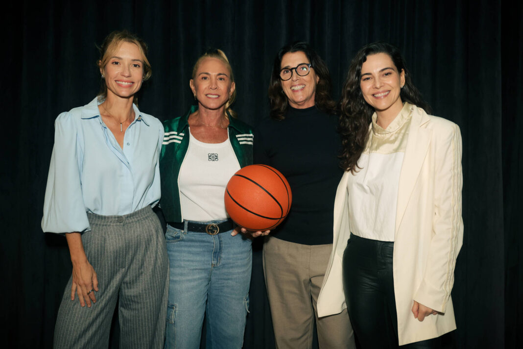 Quatro mulheres estão alinhadas em frente a um fundo preto. A segunda da esquerda segura uma bola de basquete laranja. Elas vestem roupas casuais e formais, incluindo camisa azul clara, jaqueta verde, blusa preta e blazer branco. Todas sorriem para a câmera.