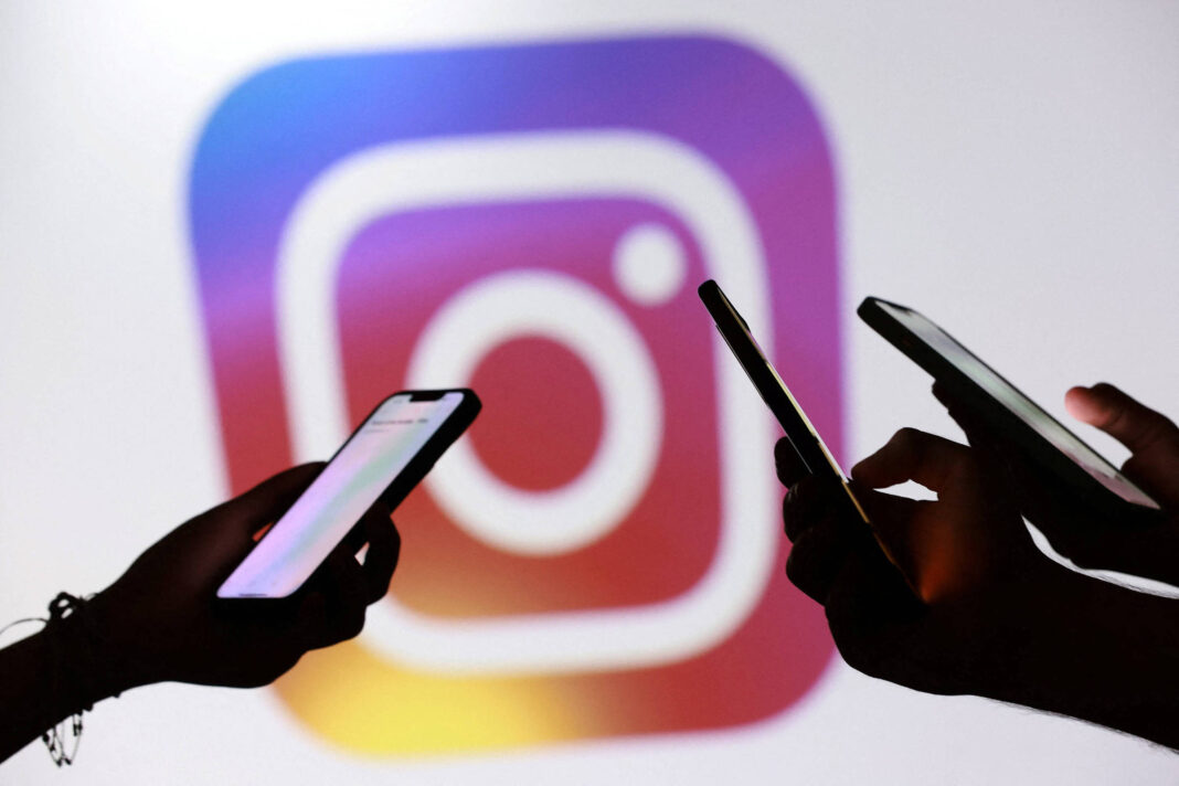 Três mãos seguram celulares com telas iluminadas em frente a um grande logo colorido do Instagram desfocado ao fundo.
