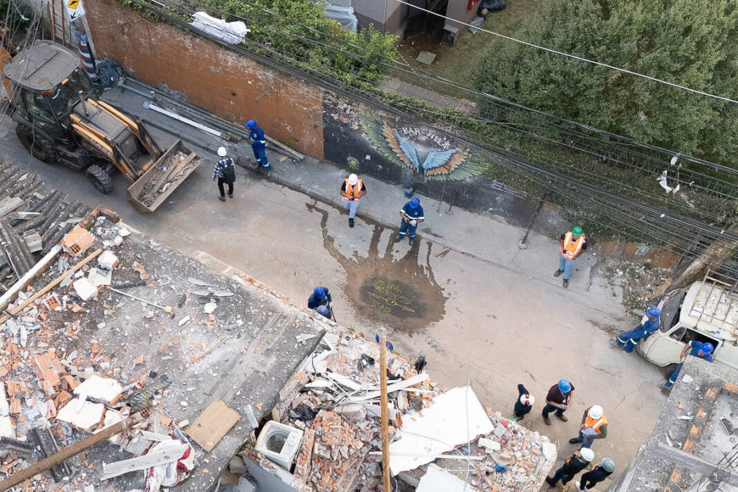 Vista aérea mostra destroços de um edifício demolido com escombros espalhados. Operários com coletes refletores e capacetes estão no entorno, junto a uma retroescavadeira estacionada na rua ao lado.