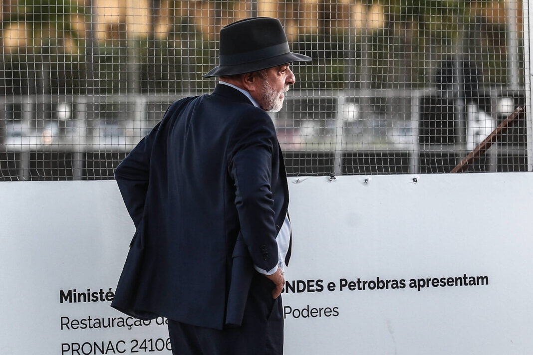 Homem de terno azul escuro e chapéu preto está de perfil olhando para uma placa branca com texto sobre restauração, atrás de uma grade metálica.