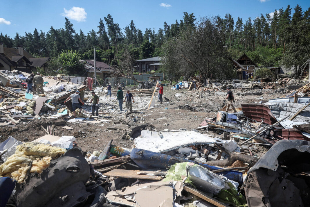 Sete pessoas recolhem destroços em terreno devastado por desastre, com casas destruídas ao fundo e árvores ao redor.