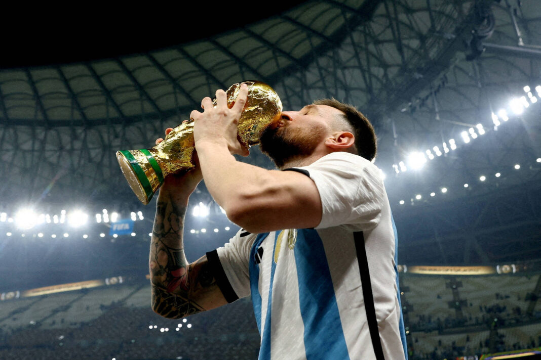 Jogador com camisa da Argentina beija a taça dourada da Copa do Mundo dentro de estádio iluminado, com arquibancadas ao fundo.