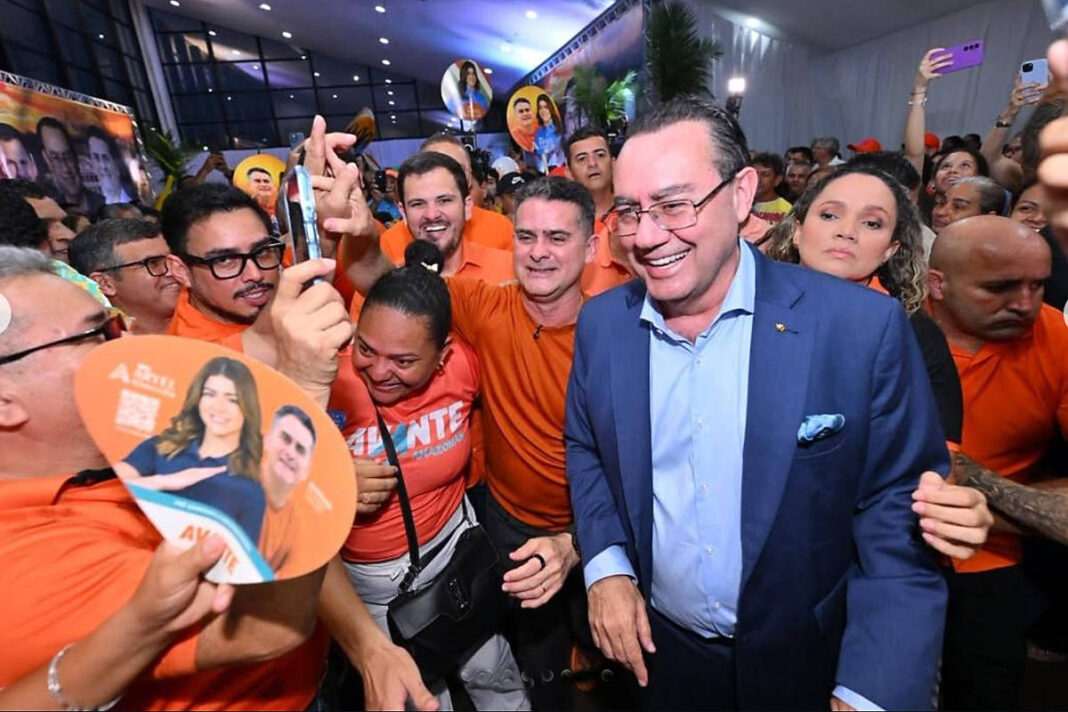 Homem de terno azul sorri enquanto é cercado por grupo de pessoas vestindo camisetas laranjas, algumas segurando cartazes e ventiladores de campanha. Ambiente interno com iluminação artificial e decoração simples.