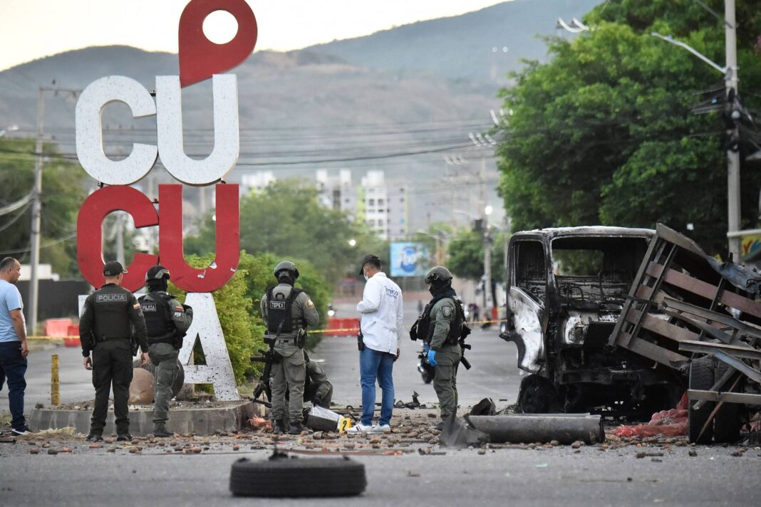 Cinco policiais e um homem de branco inspecionam rua bloqueada por destroços e veículo queimado próximo a placa grande com a palavra 