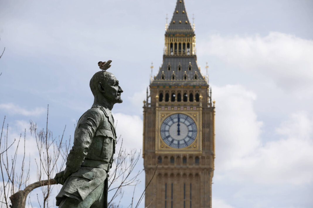 Estátua de homem em perfil com mãos atrás das costas e pássaro pousado na cabeça, com o relógio do Big Ben ao fundo em dia claro.