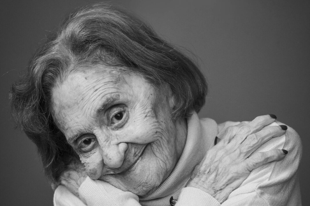Mulher idosa com cabelo curto e grisalho, vestindo suéter claro, abraça-se com os braços cruzados sobre o peito, sorrindo levemente, em fundo neutro.