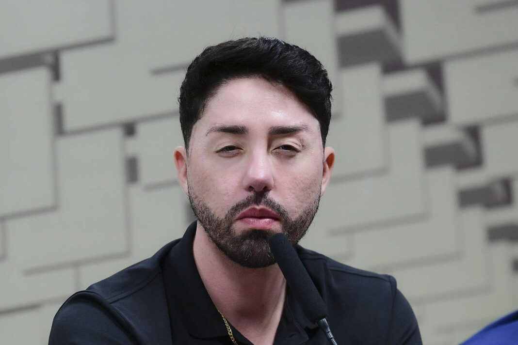 Um homem com cabelo curto e barba está sentado em uma mesa, olhando para a frente. Ele usa uma camiseta preta e está segurando um microfone. O fundo é composto por painéis em tons claros, criando um ambiente de coletiva de imprensa.