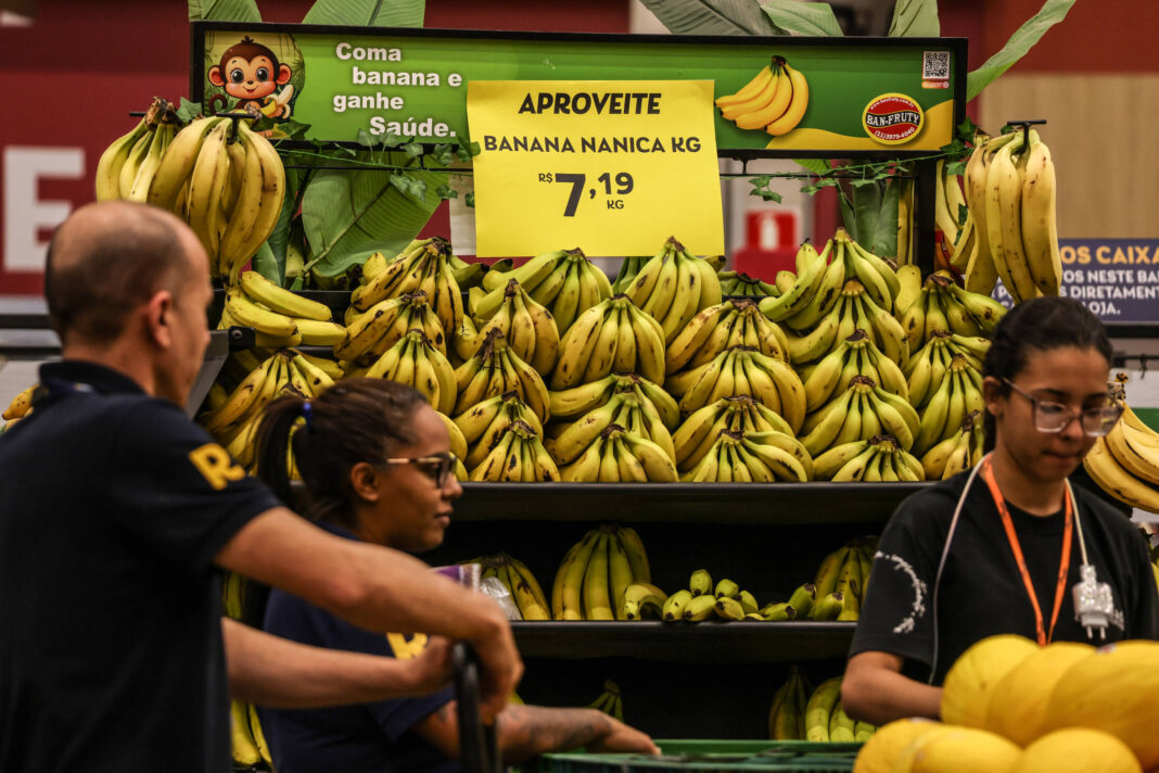 Banca de supermercado exibe várias pencas de banana nanica empilhadas em prateleiras. Placa amarela anuncia preço de R$ 7,19 por quilo. Três pessoas estão próximas à banca, duas mulheres e um homem, aparentemente clientes ou funcionários.