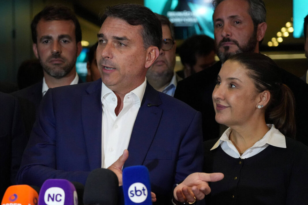 Homem de terno azul e camisa branca fala em coletiva cercado por outras pessoas. Mulher ao lado, com camisa branca e suéter preto, gesticula enquanto escuta. Microfones de emissoras estão à frente.