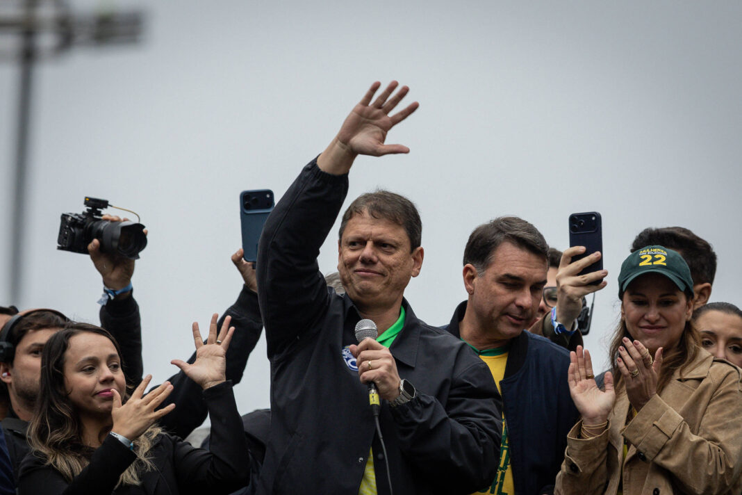 Homem no centro segura microfone e acena com a mão direita para a multidão. Pessoas ao redor filmam com celulares e aplaudem. Céu nublado ao fundo.