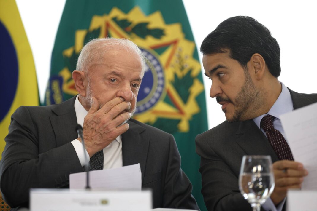 Dois homens de terno conversam em evento formal. Homem à esquerda cobre a boca com a mão enquanto ouve o outro, que segura um papel. Ao fundo, bandeira do Brasil desfocada.
