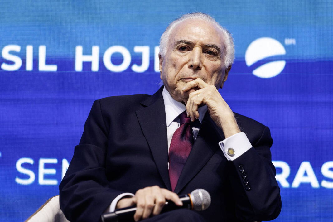 Homem idoso com cabelo branco veste terno preto, camisa branca e gravata vinho, sentado segurando microfone com uma mão e apoiando o queixo na outra, em frente a fundo azul com texto parcialmente visível.