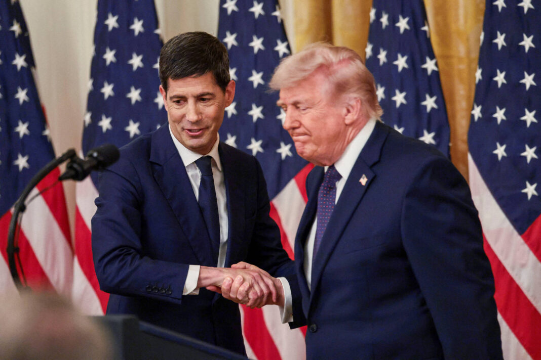 Dois homens de terno escuro apertam as mãos em frente a um conjunto de bandeiras dos Estados Unidos. O homem à esquerda tem cabelo escuro e está sorrindo levemente, enquanto o homem à direita tem cabelo loiro e expressão séria. Ambos estão em um ambiente interno com cortinas douradas ao fundo.