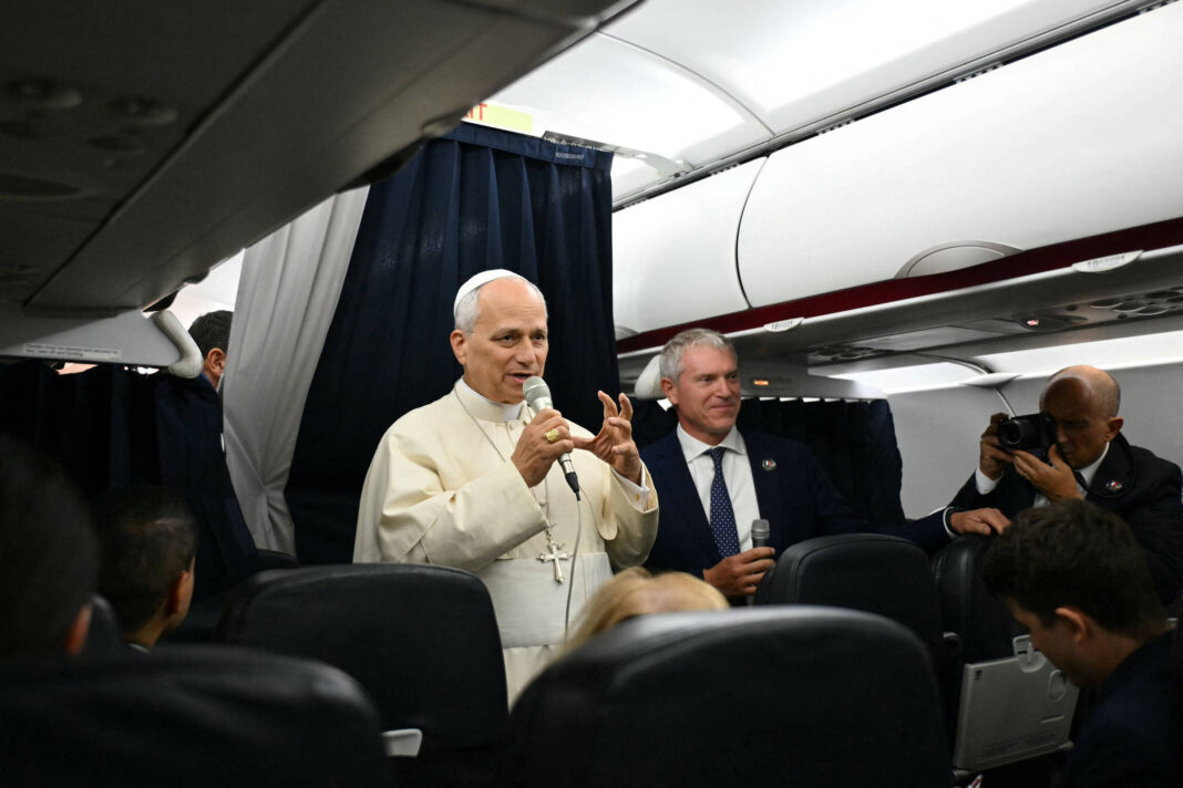Papa Francisco fala ao microfone dentro de avião, acompanhado por homem de terno segurando outro microfone. Pessoas sentadas observam, e fotógrafo registra cena ao fundo.