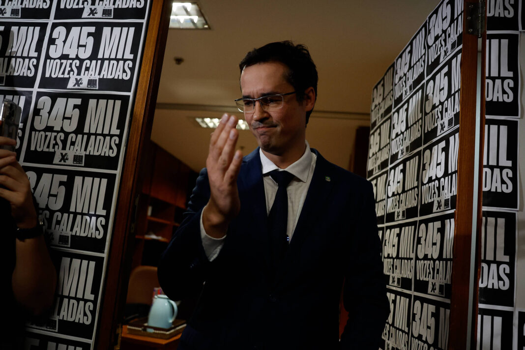Homem de terno escuro e gravata branca segura uma vela acesa com a mão direita, em um corredor estreito com painéis pretos e brancos ao fundo contendo textos repetidos.
