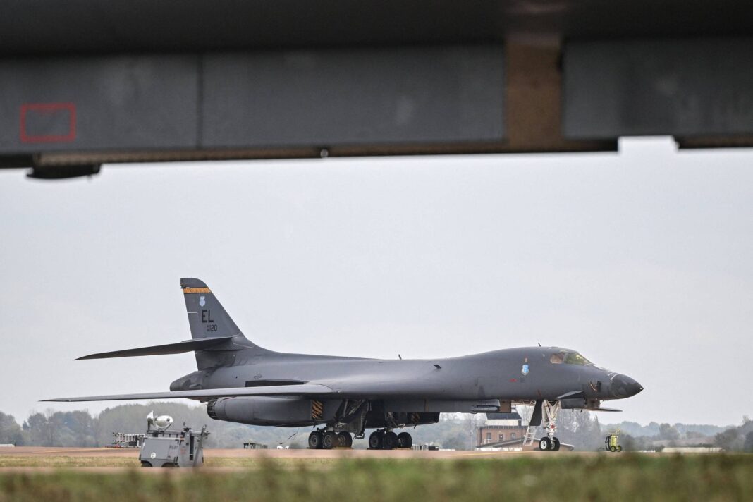 Avião militar B-1B estacionado na pista de aeroporto sob estrutura metálica. Veículo de apoio branco está próximo à frente da aeronave. Céu nublado e área de grama ao redor da pista.