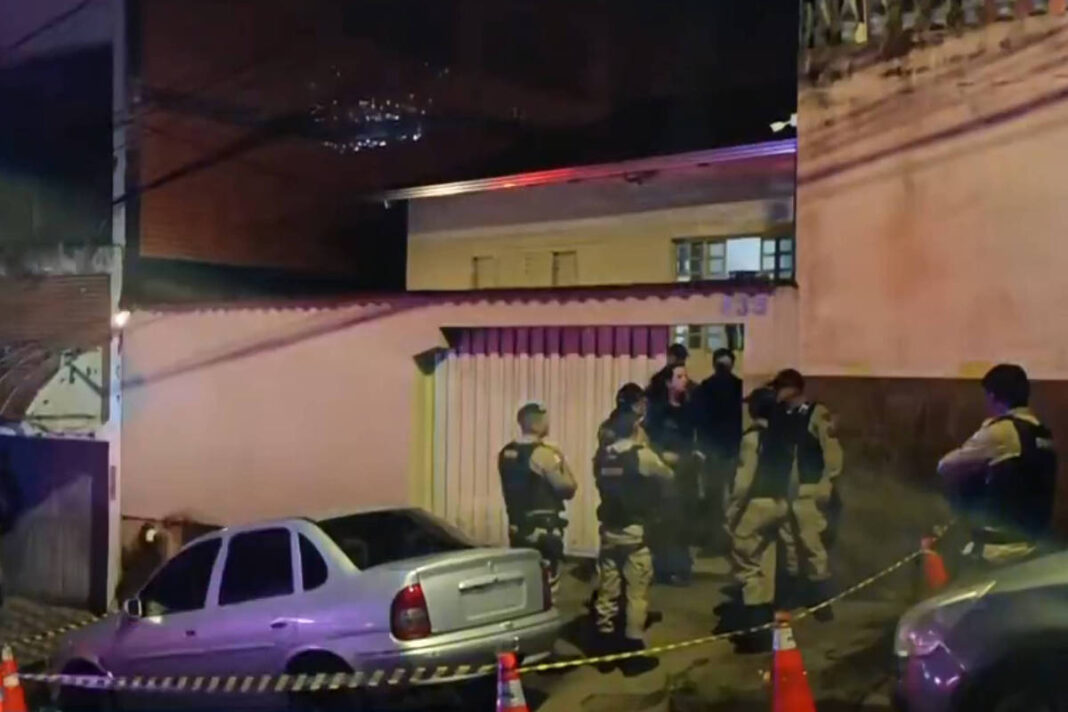 Seis policiais uniformizados estão reunidos em frente a um portão de garagem em uma rua residencial à noite. Dois carros estão estacionados próximos, e a área está isolada com cones laranja e fita de segurança.